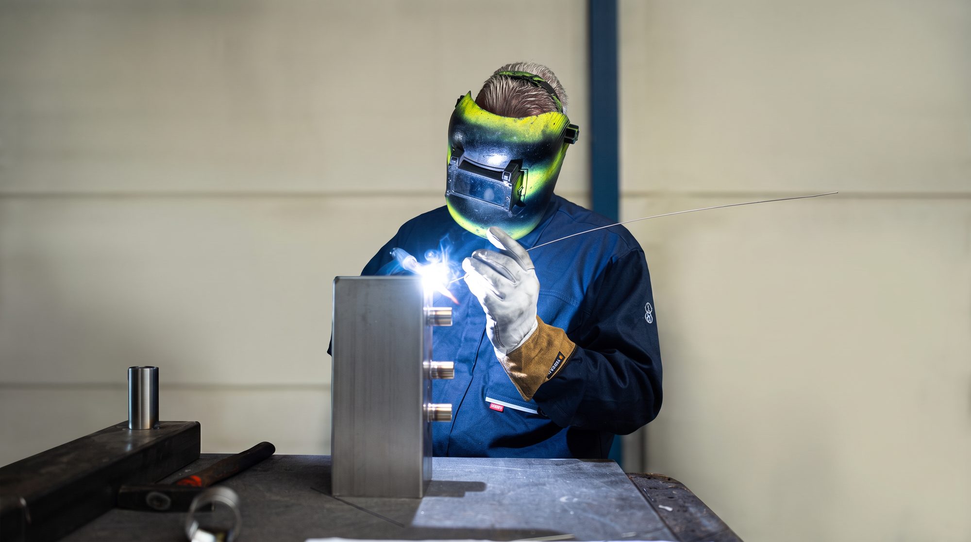 TIG-lasser in blauwe overall met groen lasmasker werkt aan een roestvaststalen werkstuk
